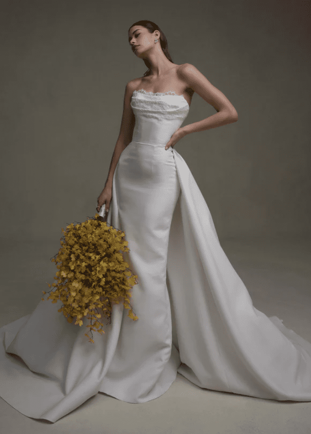 1745069798876-screen-shot-2025-04-19-at-9-36-09-am Senstudio's ivory fit-and-flare wedding gown (model SE070), featuring a strapless bodice with delicate lace detailing on the upper edge and crafted from luxurious matte mikado. The fitted skirt expands into a dramatic detachable overskirt that forms a sweeping cathedral train. A model poses holding a large, cascading bouquet of bright yellow flowers.