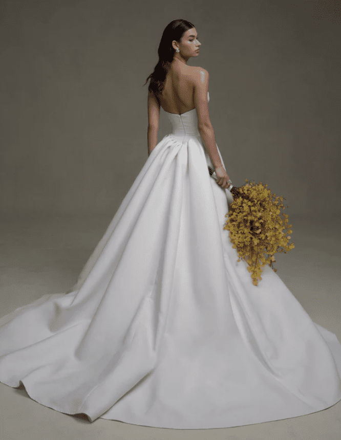 1745069798876-screen-shot-2025-04-19-at-9-36-19-am SE070, an ivory strapless fit-and-flare wedding gown by Senstudio, crafted in luxurious matte mikado. The model is viewed from the back, looking over her right shoulder, holding a large cascading yellow floral bouquet. The gown features a dramatic detachable overskirt, creating a full silhouette that flows into a sweeping cathedral train, with delicate lace detailing across the front of the upper bodice.