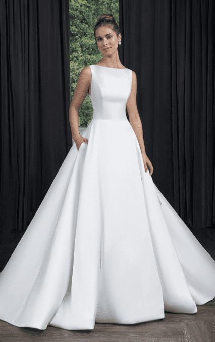 1745250261839-screen-shot-2025-03-20-at-4-39-05-pm 55086, an Ivory refined Mikado ball gown, is worn by a model with an updo and pearl earrings, posing with one hand in a pocket against a dark curtain backdrop with greenery visible. This timelessly sophisticated dress features an elegant bateau neckline and princess seams on the bodice, creating a structured, regal silhouette. It includes a striking square back, a dramatic detachable Mikado bow, Mikado-covered buttons trailing to the end of the chapel-length train, and a horsehair hem ensuring its graceful shape and volume.