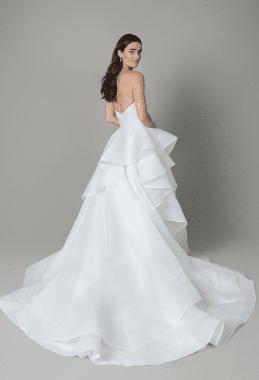 1745250485223-screen-shot-2025-04-21-at-11-47-11-am 55210, an ivory strapless organdy ball gown, shown from the back with a model looking over her shoulder. It features a pleated bodice with an asymmetric neckline, a voluminous ruffle skirt with horsehair-edged layers, and a chapel-length train. Organdy-covered buttons run down the back of the bodice.