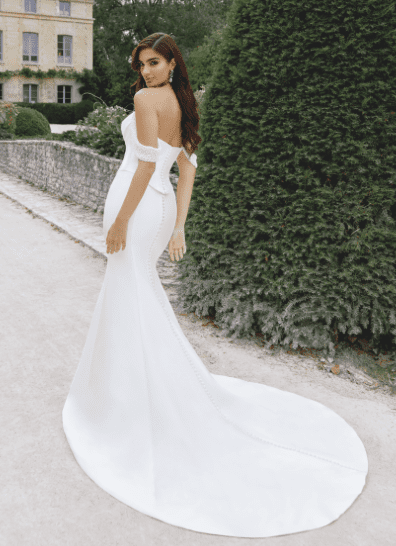 1745251014976-screen-shot-2025-04-21-at-11-56-00-am An ivory mikado two-piece fit-and-flare wedding gown, featuring a draped off-the-shoulder sweetheart bodice with delicate pearl beading and a structured corset. The skirt has covered buttons extending down the back to a chapel-length train. A model with long dark hair stands on an outdoor path, looking back over her shoulder, with a stone wall, green foliage, and a historic building in the background.
