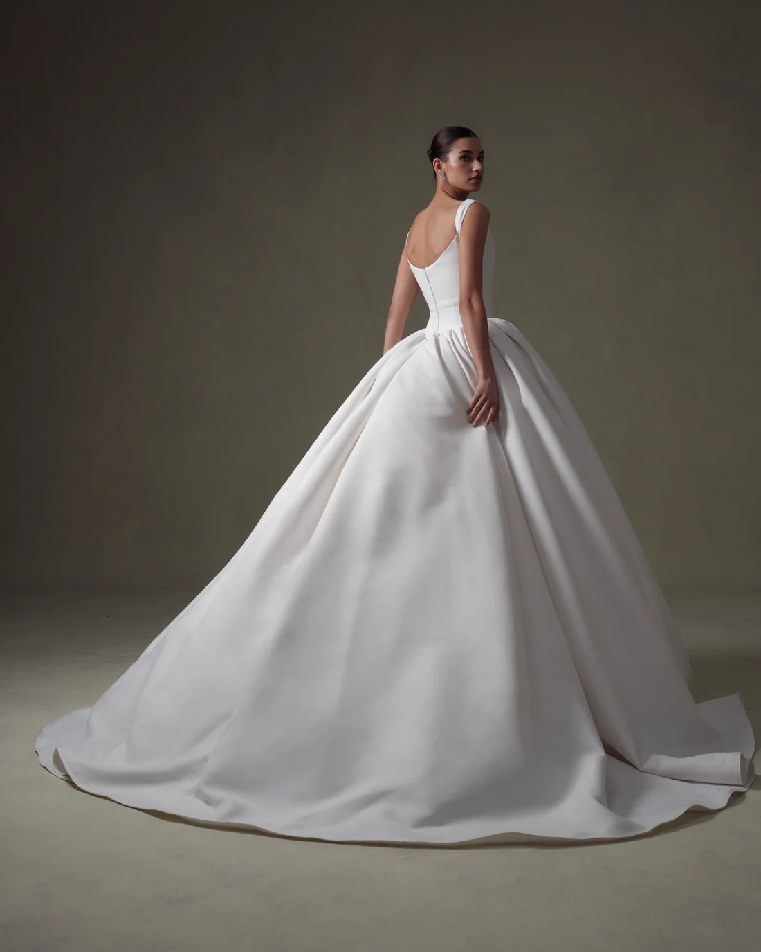 1745434593453-20250313NF4916 SE067, an ivory matte Mikado ball gown by Senstudio. A model is shown from the back, looking back over her left shoulder, highlighting the gown's structured bodice with elegant straps, a modern basque waist, and its voluminous full skirt with a dramatic chapel-length train, showcasing a clean, regal look.