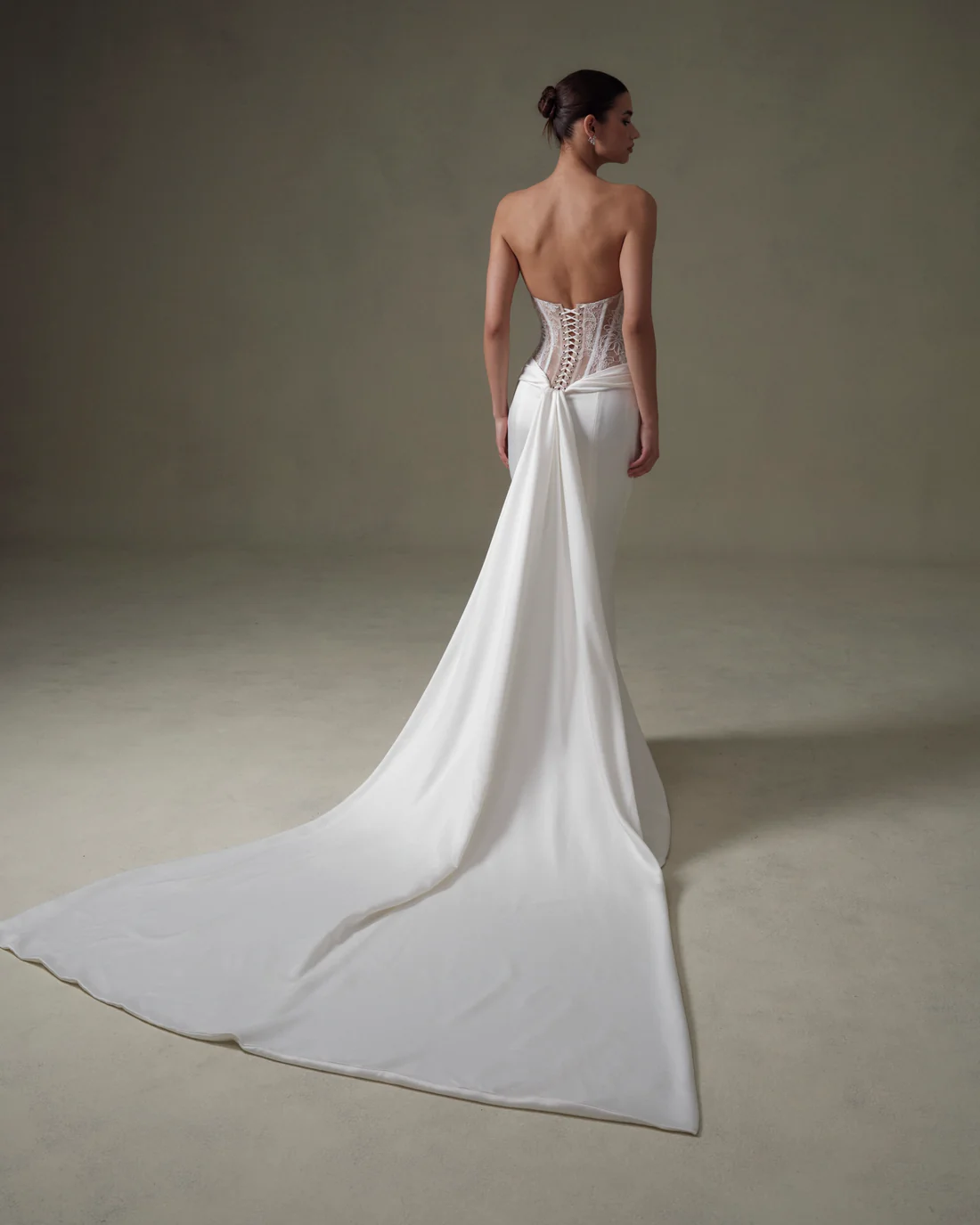 1745435042284-20250313NF4806 An ivory and nude strapless mermaid wedding gown from Senstudio, modeled from the back. The dress features a sculpted, unlined lace corset bodice with crisscross lacing, leading to a sleek ivory skirt that flares into a dramatic panel train flowing behind the model.