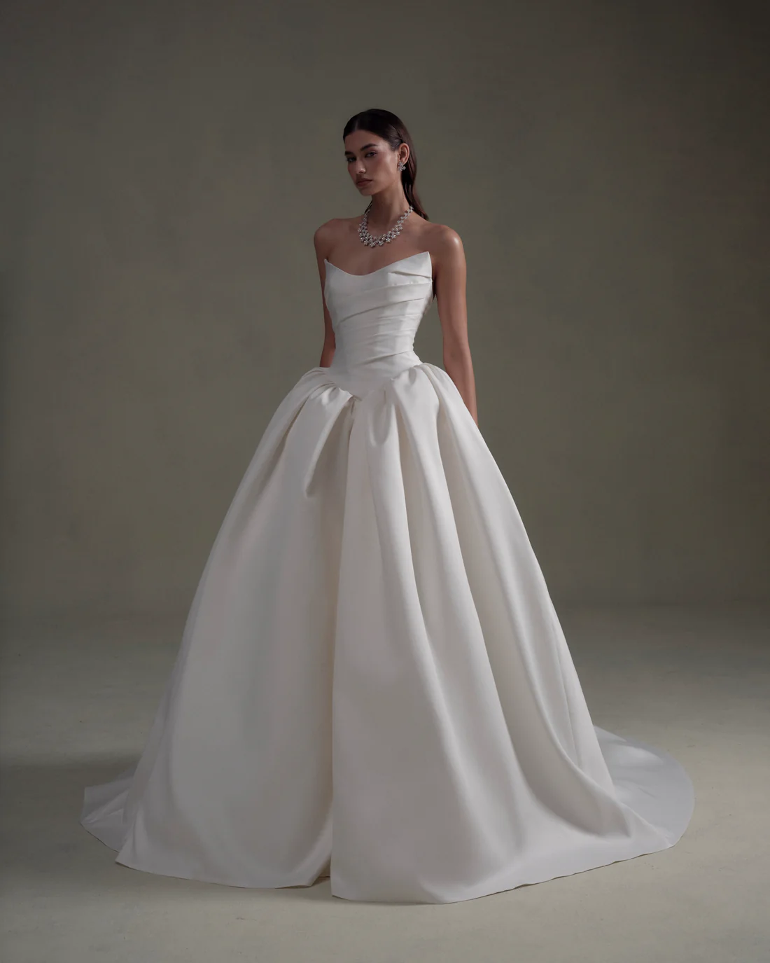 1745436421871-20250313NF3822-1 SE086, an ivory strapless bridal ball gown by Senstudio, featuring a unique cateye neckline and a softly draped bodice. The dress has a voluminous, pleated skirt that dramatically cascades to the floor with a cathedral-length train. A model wears the gown and a sparkling diamond necklace.
