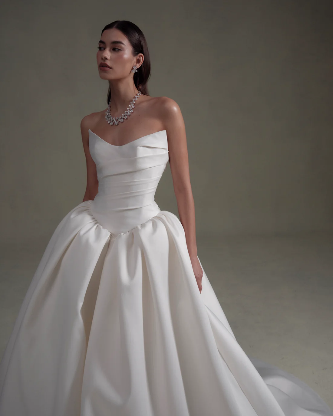 1745436421872-20250313NF3841 SE086, an ivory strapless ball gown featuring a unique cateye neckline, a draped bodice, and a voluminous pleated skirt with a cathedral train. The gown is modeled by a woman with dark hair, wearing diamond earrings and a necklace.