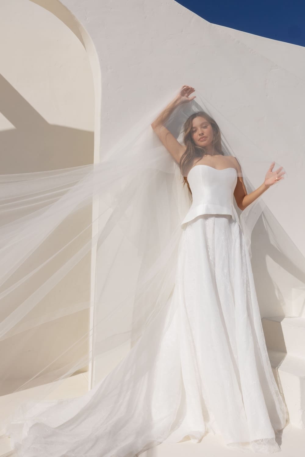 1745441292967-3HTC23gg Sofia, an ivory two-piece bridal gown featuring a structured Mikado strapless sweetheart bodice cinching the waist and an airy A-line skirt of lace and organza. A model, partially veiled by a flowing cathedral veil, poses against a bright white wall with architectural details and a clear blue sky.