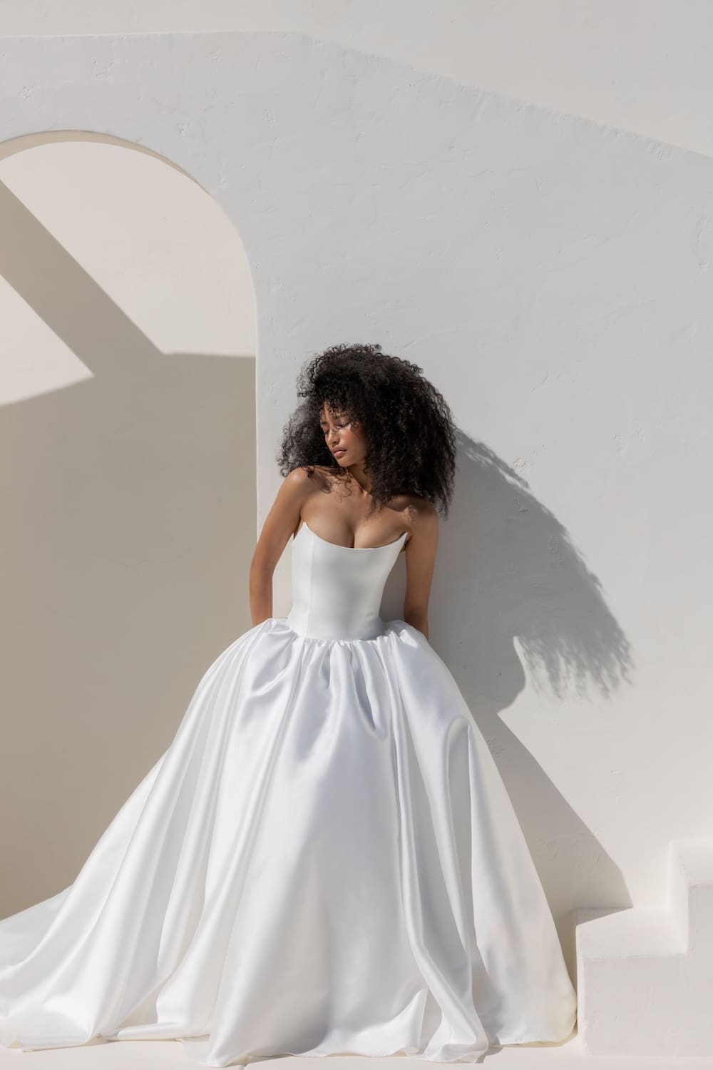 1745441511043-BJr4c_vA Lucinda, an ivory strapless ball gown with a refined scoop neckline and a Basque waist, crafted from lustrous Italian Mikado, featuring dramatic volume, worn by a model with voluminous dark curly hair, leaning against a sunlit white wall with an architectural archway.
