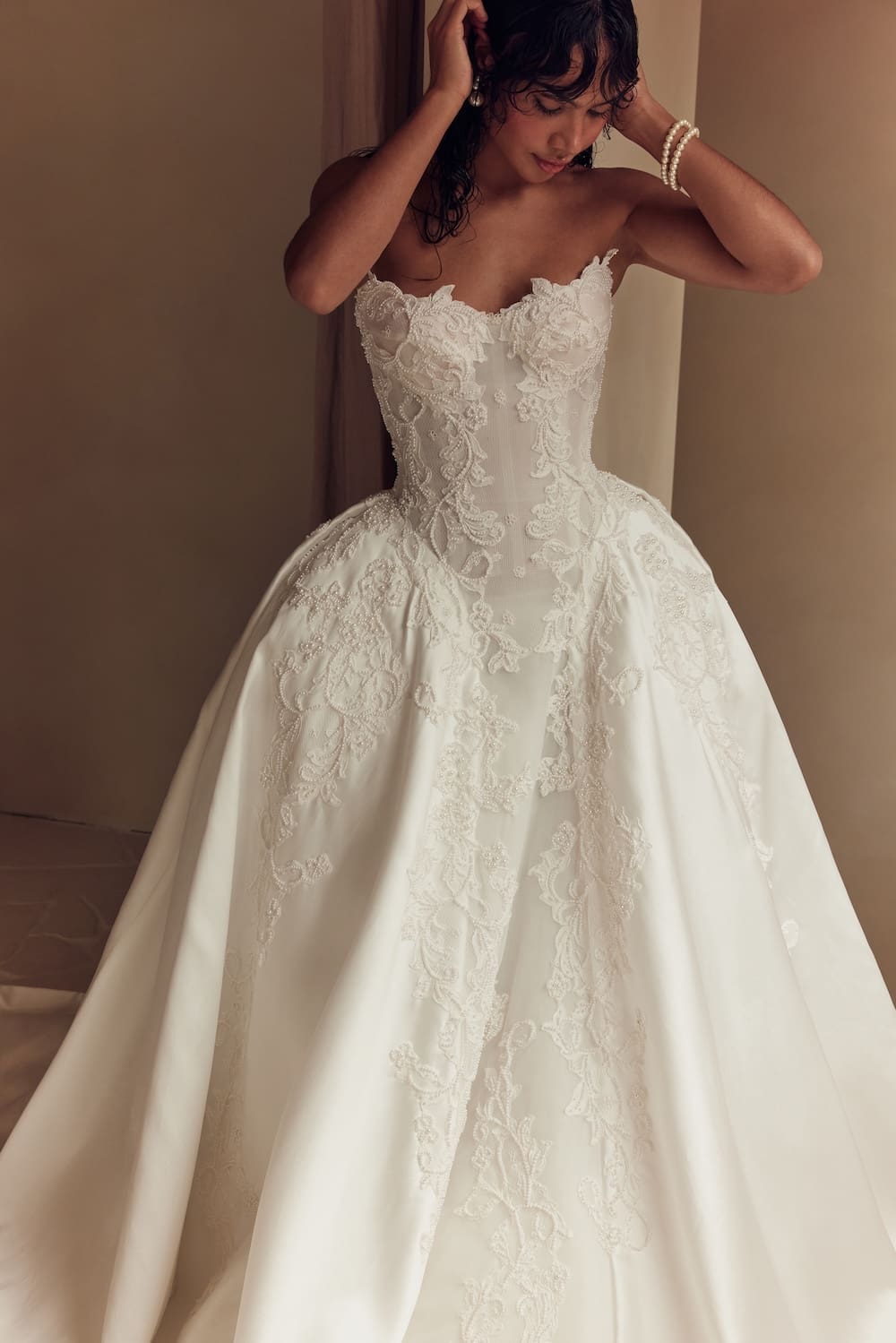 1747162862887-leah-da-gloria_09_031 Alexandra, an ivory strapless bridal ball gown by Leah Da Gloria, features a sweetheart neckline, a fitted Basque-waist bodice, and a voluminous mikado skirt, all richly embellished with intricate lace and shimmering beaded embroidery. A woman with dark, wet-look hair, adorned with pearl earrings and a pearl bracelet, gazes downward while posing with her hands near her head, showcasing the elegant gown.
