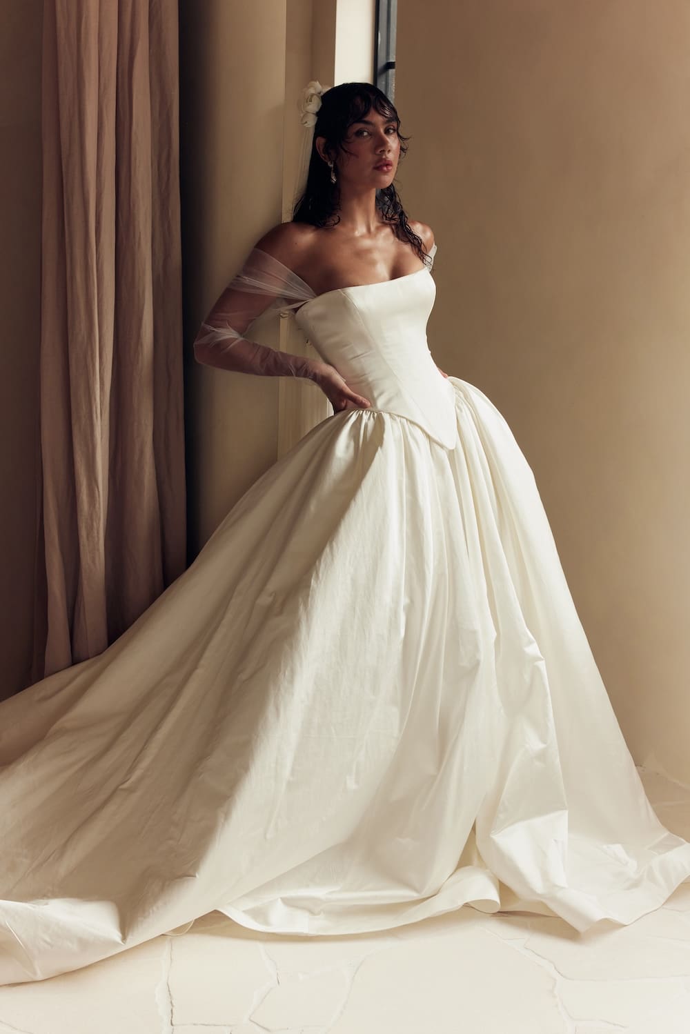 1747163533273-leah-da-gloria-03-042 Elizabeth, a Natural White strapless satin ballgown featuring a structured Basque waist, a full, dramatic skirt, and soft, off-the-shoulder tulle sleeves. The model, with dark, wet hair and bangs, looks intently at the viewer while posing elegantly against a warm-toned wall with curtains.