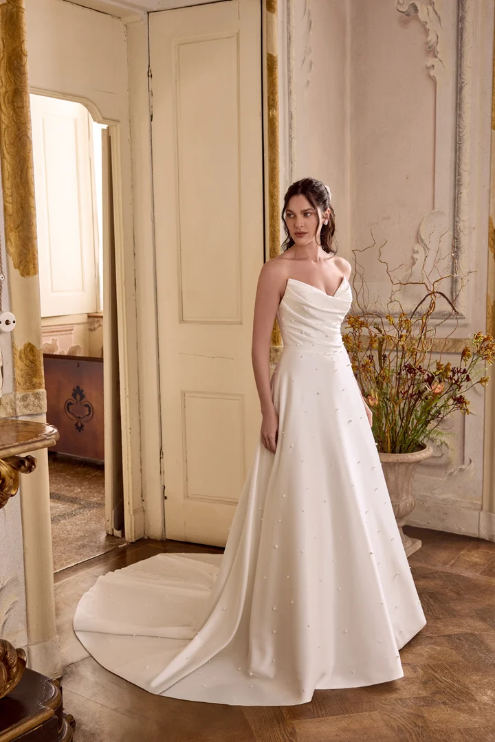1747243336690-88475-lizzie-ff-justin-alexander-2-1 Lizzie, an ivory A-line wedding gown by Justin Alexander, crafted in luxurious charmeuse. It features a pointed sweetheart neckline, soft ruching across the bodice, and scattered pearl embellishments throughout the bodice and A-line skirt. The gown also has hidden pockets and is finished with buttons trailing to the end of its chapel-length train.