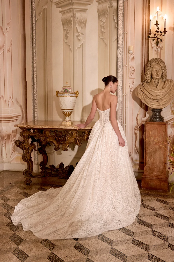 1747243543708-88485-love-jab-ss26-fb-justin-alexander-1 Love, a Sand/Ivory/Latte strapless bridal ball gown by Justin Alexander. The gown features a demi-sheer sweetheart bodice with exposed boning, a flattering Basque waist, side gathering on the voluminous skirt, and a chapel-length train. It's crafted from allover beaded and sequined lace, accented with crystals and pearls, and includes a row of buttons at the back waist and a structured horsehair hem. The model, viewed from the back, stands gracefully in an ornate hall, her hand resting on a carved marble table, highlighting the gown's luxurious train.