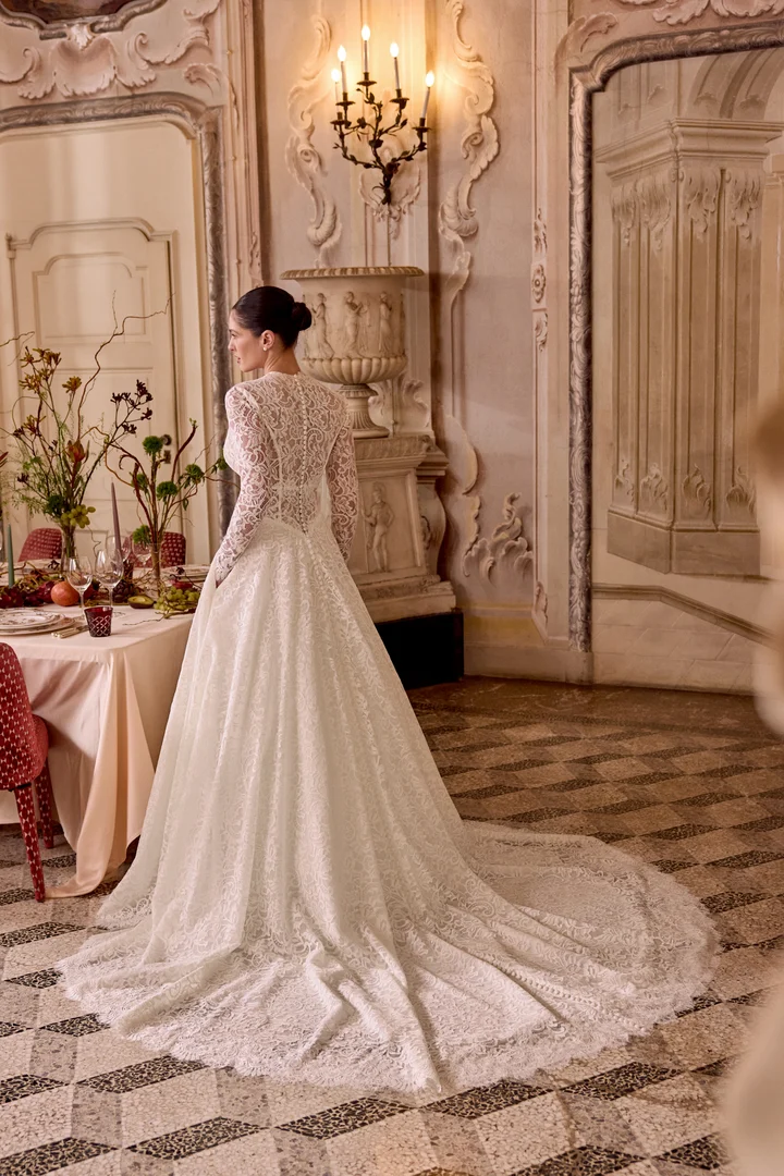 1747243620359-88477-lady-fb-justin-alexander-5 Lady, an Ivory/Latte A-line bridal gown by Justin Alexander, featuring allover lace, long sleeves with scalloped cuffs, a Basque waist, and a high illusion back with buttons cascading to a chapel-length train. The bride is seen from behind in an ornate room, looking left towards an elegantly set table with flowers.