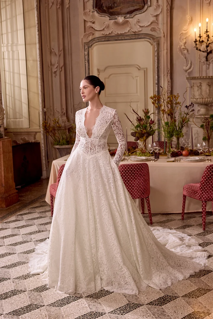 1747243620360-88477_Lady_FF_Justin-Alexander_1 Lady, an ivory and latte A-line wedding gown crafted from allover lace, features a plunging V-neckline with scalloped lace, a demi-sheer bodice with a Basque waist, and long illusion lace sleeves with scalloped cuffs. The dress boasts a chapel-length train. A model with her hair in a sleek bun poses elegantly in a grand, ornately decorated room with a patterned floor and a beautifully set dining table visible in the background.