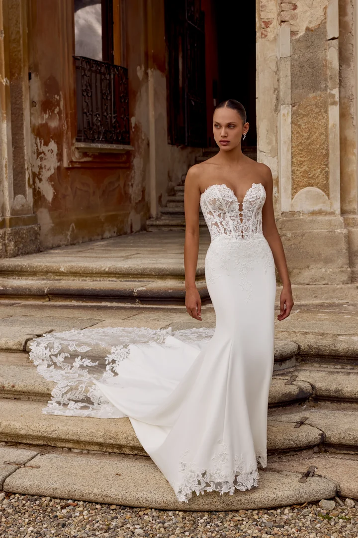 1747243789745-88353_Hope_FF_D_2025_RL_Justin-Alexander_2 Hope, an ivory and latte stretch crepe fit-and-flare bridal gown, features a plunging sweetheart neckline with exposed boning and sequined lace appliqués on the bodice, creating a sculpted Basque waist. The gown's skirt has a striking cutout hem with lace appliqués that extend into a dramatic cutout cathedral train. The gown is worn by a model with dark hair pulled back, standing on weathered stone steps in front of an old, ornate building.