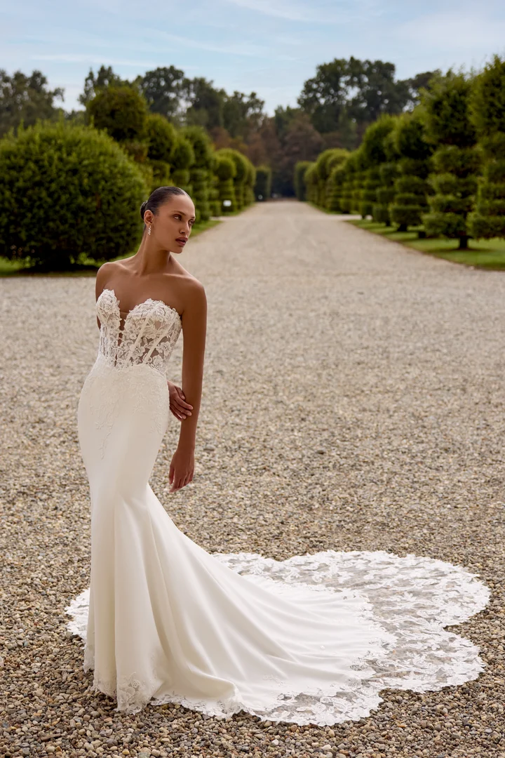 1747243789745-88353_Hope_FF_D_2025_RL_Justin-Alexander_3 Hope, an Ivory/Ivory/Latte stretch crepe fit-and-flare bridal gown, features a plunging sweetheart neckline with exposed boning and a Basque waist. Sequined lace appliqués adorn the bodice, extend down the skirt, and form a striking cutout hem that flows into a dramatic cutout cathedral train. The gown is shown on a model with dark hair pulled back, standing on a gravel path with a tree-lined avenue in the background.