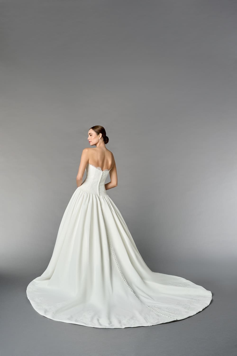 1747244950742-rng-55269-fb Iris, an Off White/Ivory satin ballgown, features a strapless modified sweetheart neckline framed by delicate scalloped lace which also traces the elegant inverted dropped waist. The bodice is richly layered with Chantilly and corded Alençon lace, leading to a full, gently gathered satin skirt. A row of buttons runs down the chapel-length train, which is embellished with lace appliqués at the hem. The model is shown from the back, looking over her left shoulder.