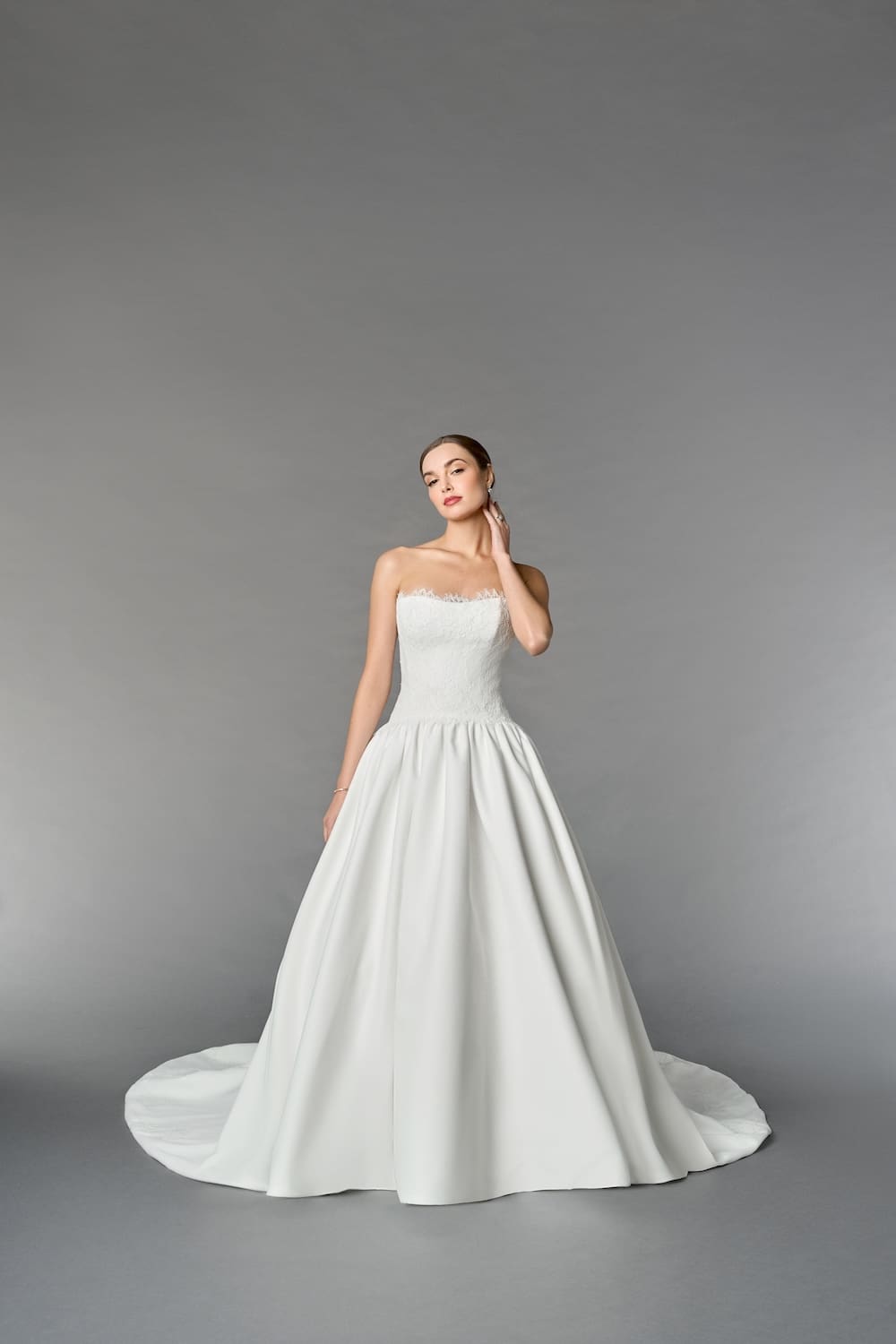 1747244950744-RNG_55269 Iris, an Off White/Ivory satin ballgown bridal gown, featuring a strapless modified sweetheart neckline with delicate scalloped lace tracing the neckline and inverted dropped waist. The bodice showcases layered Chantilly and corded Alençon lace, while the full satin skirt has gentle gathers and discreet pockets. The gown is completed with a row of buttons down its chapel-length train and lace appliqués embellishing the hem. A model with fair skin and hair pulled back poses, looking towards the viewer, with one hand elegantly touching her neck.