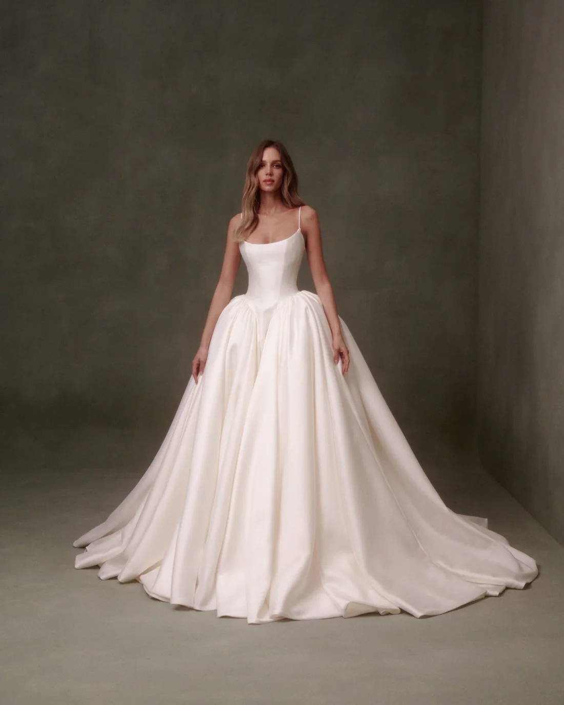 1748022352352-2024091132769 NF095, a white Nicole + Felicia textured Mikado ballgown, featuring a scoop neckline with delicate spaghetti straps, a sculpted Basque waist, and a voluminous skirt that flows into a cathedral train. The structured yet romantic gown is worn by a model standing against a dark olive green background.