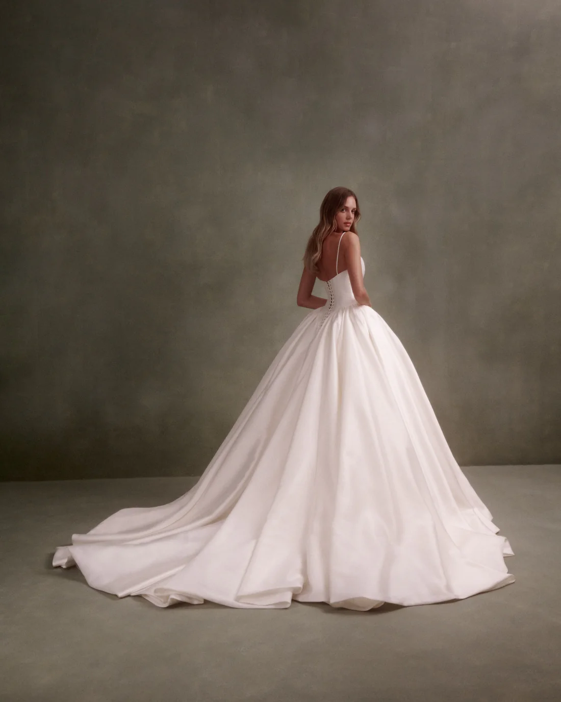 1748022352353-2024091132804_b813116d-2244-4bbb-9e61-339276089a01 NF095, a beige textured Mikado ballgown by Nicole + Felicia, featuring a scoop neckline, delicate spaghetti straps, a Basque waist, and a dramatic lace-up corset back. The gown's voluminous skirt and cathedral-length train are showcased as the model, with long blonde hair, looks over her right shoulder while standing against a muted green studio backdrop.