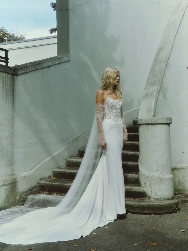 1749144031736-rv-fw25-rivini-campaign-sennaf Senna, an ivory Rivini bridal gown, is modeled by a blonde woman standing on a concrete path next to worn stone stairs and a light wall. The sleek crepe sheath gown features a flattering Basque waist and a translucent boned bodice adorned with intricate floral beaded lace, complemented by a modern cat-eye neckline. She wears soft, ruched tulle detachable sleeves, and a dramatic, ethereal English tulle choker-cape trails behind her, forming a cathedral-length train on the steps.
