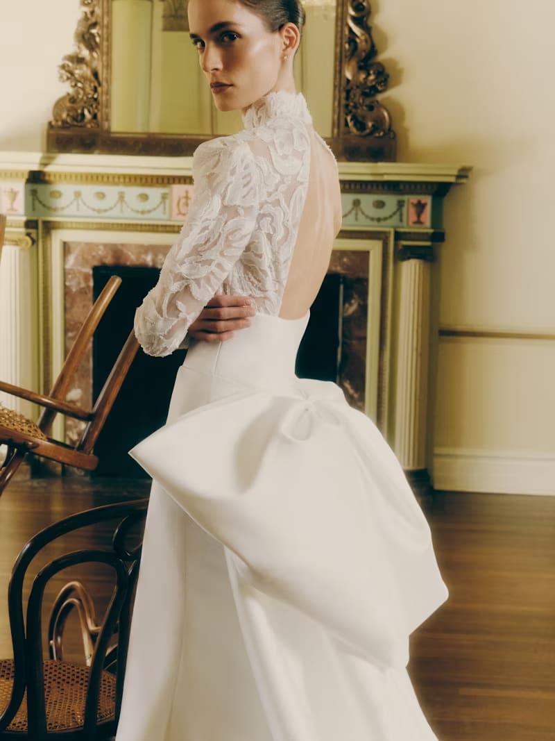 1749144200158-rv_ss26_rivini_kit-1 Kit, an ivory fit-and-flare bridal gown in luminous Neo-Satin, features a sheer floral lace bodice with long sleeves, a refined mock neckline, and an elegant keyhole back. A detachable pleated overskirt with a couture bow adds drama to the back.