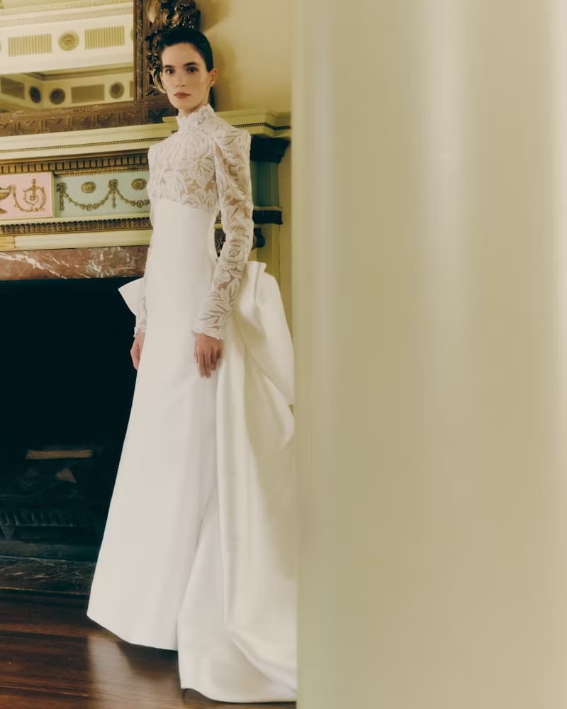 1749144200159-rv-ss26-rivini-kit-3-front-1 Kit, an ivory fit-and-flare wedding gown by Rivini, featuring a striking demi-sheer floral lace bodice with long sleeves and a high mock neckline, a banded empire waist, and a luminous satin skirt with a dramatic detachable pleated overskirt and couture bow. The gown is modeled by a woman standing elegantly indoors beside an ornate fireplace.