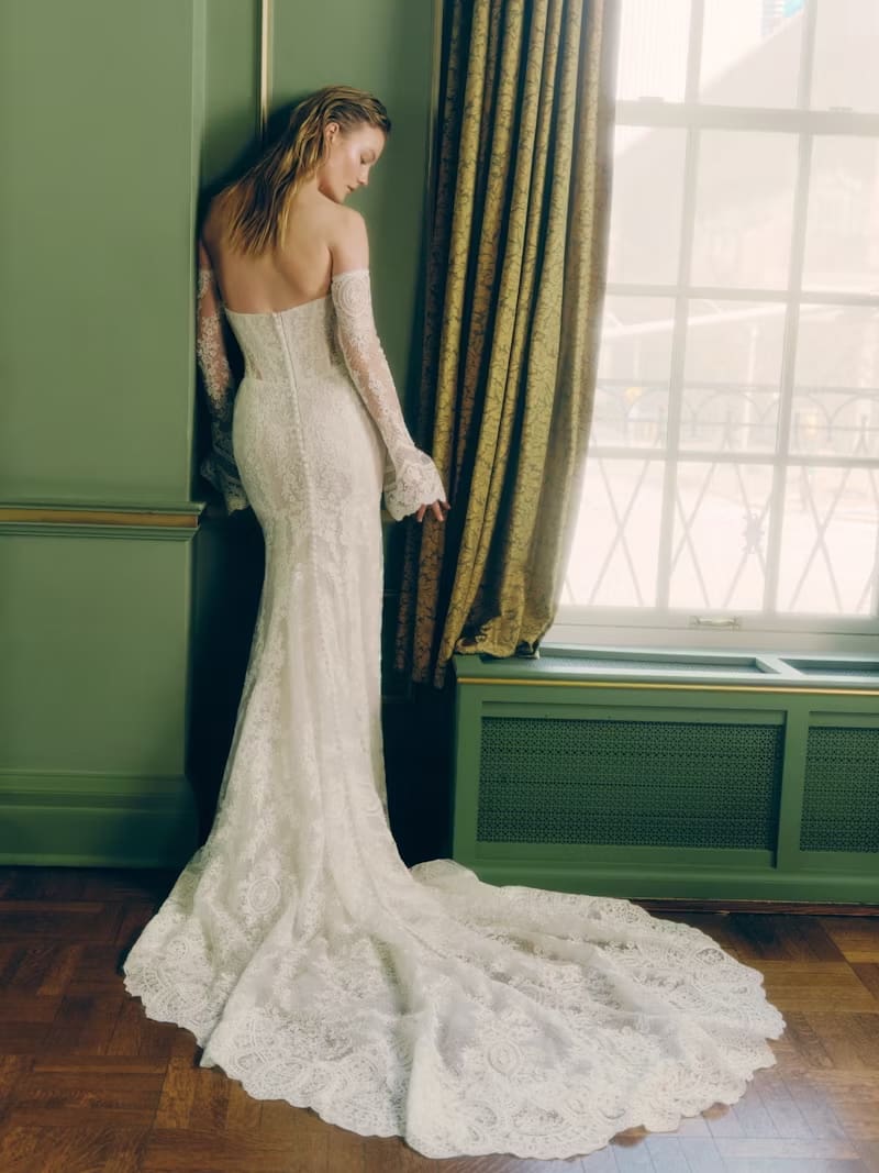 1749144305228-rv-ss26-rivini-viv-3-back Viv, an ivory and nude lace fit-and-flare bridal gown by Rivini, featuring a translucent sheer corset bodice adorned with hand-appliquéd lace, elegant detachable lace opera cuffs, and a row of buttons trailing down the back to its chapel train.