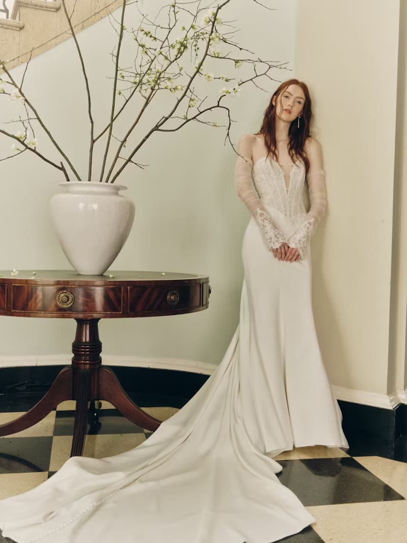 1749145344931-rv-ss26-alyne-jenna-1-front-copy Jenna, an ivory sheath wedding gown by Alyne, featuring a plunging sweetheart neckline, a translucent lace bodice with a modern corset-inspired design, and a flattering Basque waist. The sleek crepe skirt flows into a cathedral train, complemented by romantic removable lace opera cuffs and delicate buttons down the back. The gown is modeled by a woman with long red hair, standing next to a dark wooden table adorned with a large vase of flowering branches, against a light wall and a black and white checkered floor.