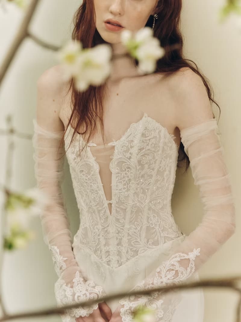 1749145344932-rv-ss26-alyne-jenna-3-copy Jenna, an ivory sheath bridal gown by Alyne. This striking design features a plunging sweetheart neckline and a flattering Basque waist. The translucent lace bodice showcases exposed back boning for a modern, corset-inspired silhouette, with delicate buttons trailing down the back to a cathedral hem. Romantic lace opera cuffs adorn the removable sheer sleeves. Available in size 12. Additional attributes include: Fit n Flare, Strapless, Crepe.