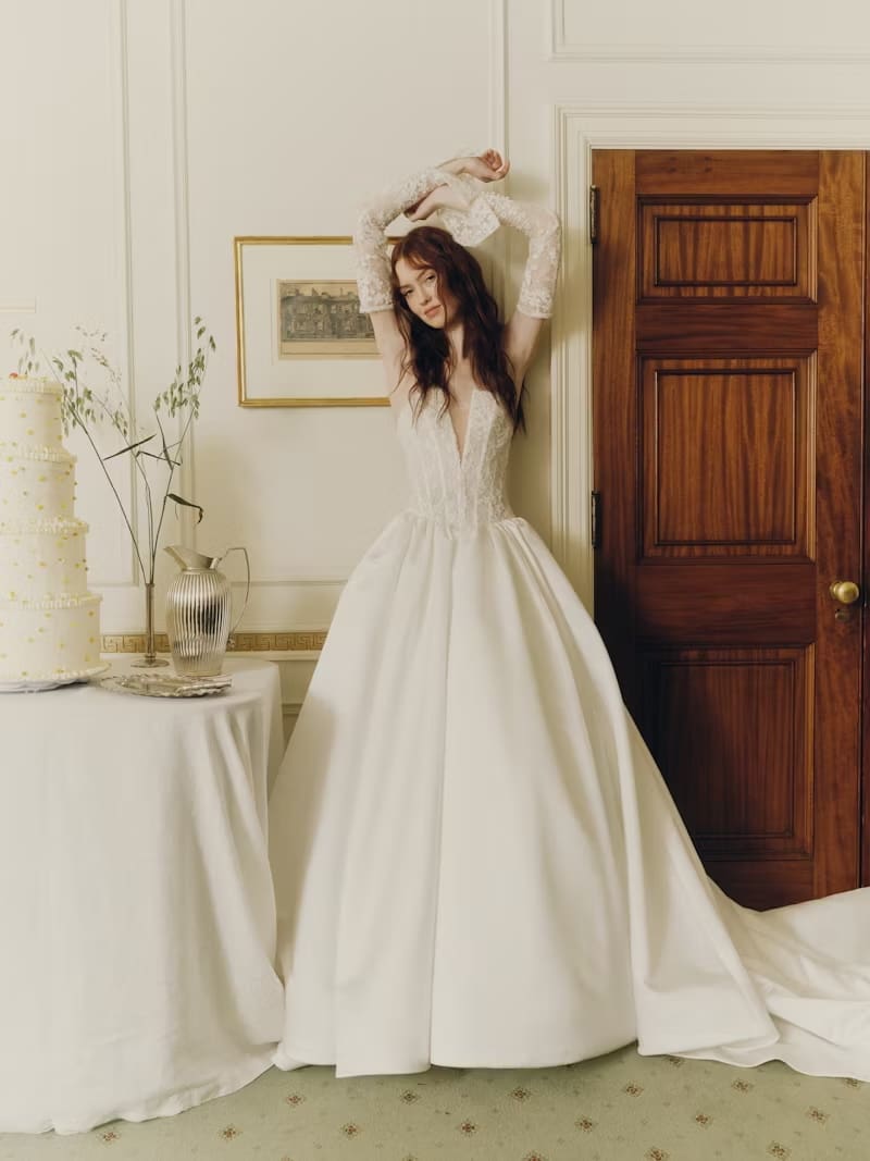 1749145775003-rv-ss26-alyne-mabel-1-front-1 Majenna, an Off White strapless ballgown featuring a translucent beaded lace bodice with a plunging V neckline, paired with a voluminous satin skirt that creates a dramatic silhouette and flows into a cathedral train. Pearl buttons trail elegantly down the back of the skirt. The model, with long reddish-brown hair, wears delicate lace opera cuffs (removable sleeves) and strikes a pose with her arms raised overhead, leaning against a cream paneled wall in a formal room. A multi-tiered wedding cake and silver serving ware are visible on a draped table to the left, with a dark wooden door on the right.