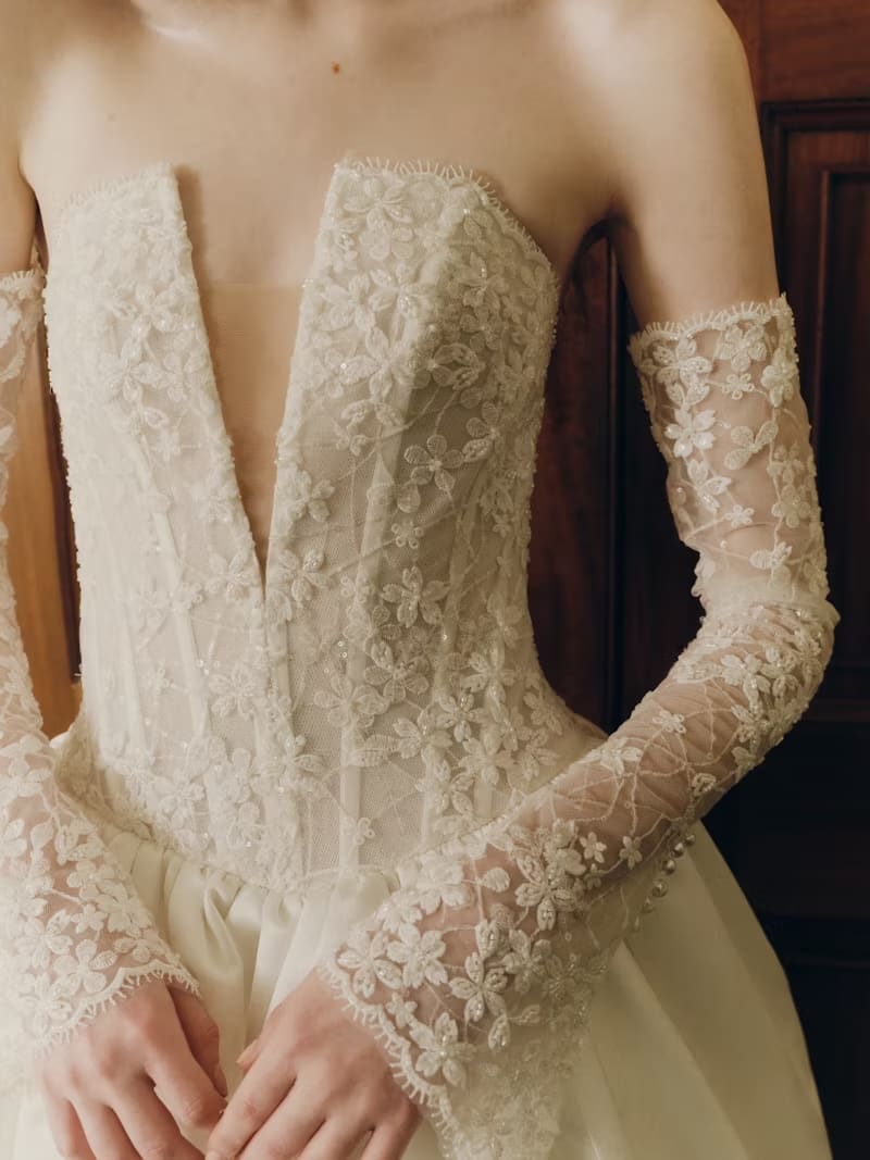 1749145775004-rv-ss26-alyne-mabel-2 Majenna, an off white strapless ballgown by Alyne, featuring a translucent beaded lace bodice with intricate floral patterns, sequins, and beads, and a plunging V neckline. Delicate lace opera cuffs (removable sleeves) with matching floral embroidery adorn the model's arms. A voluminous satin skirt is partially visible at the bottom.