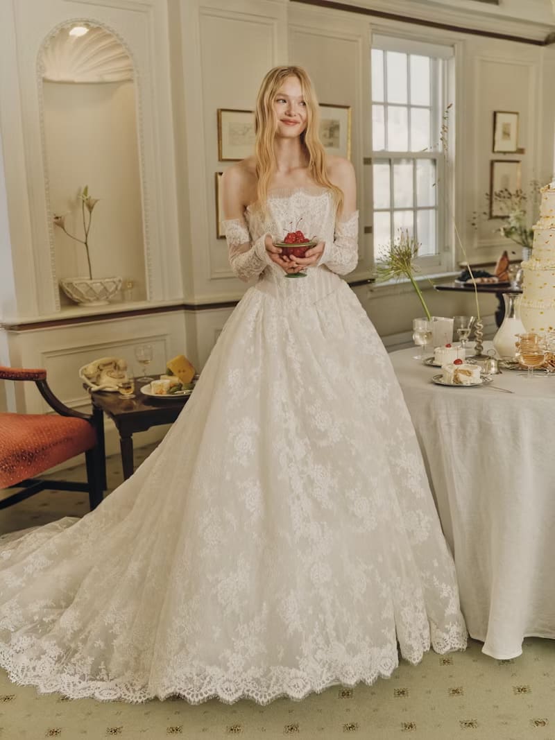 1749147134186-rv-ss26-alyne-dawes-2-front-1 Dawes, an ivory strapless lace Basque waist ballgown by Alyne, is worn by a smiling blonde model. The bridal gown features a demi-sheer lace bodice with micro-pleated tulle at the neckline, a dramatic diaphanous lace skirt with a cathedral-length train and buttons trailing down the back, and elegant removable lace opera cuffs. She holds a small bowl of cherries and stands in an elegant room with a tiered cake and refreshments.