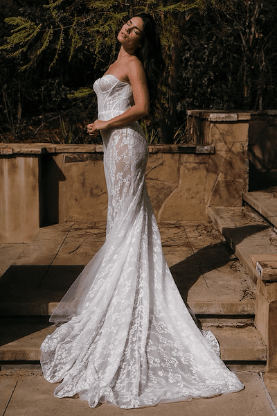 1756319453763-0baeb4_baceac35e0794efd90b5f0bd569266dd-mv2 Liana, an ivory and ivory mermaid wedding gown by Reve The Label, features a dramatic fit-and-flare silhouette and cathedral train. It has a structured, strapless corset bodice with a sweetheart neckline, crafted from a luxurious blend of floral French lace and sheer tulle. The gown beautifully accentuates the body with sheer panels, creating a modern, barely-there effect visible through the intricate lace. The model is pictured outdoors, basking in sunlight against a textured stone wall and green foliage, head tilted back with eyes closed.