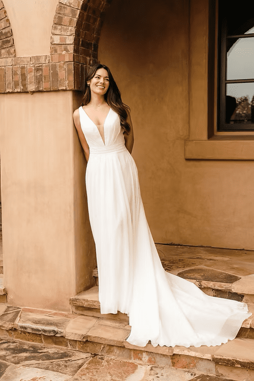1756319968327-0baeb4_4a1d2e52129a4b03bc9a9c0ac4522bc6-1 Noemi, an ivory chiffon A-line wedding gown, worn by a smiling model leaning against a textured brown wall. The dress features a plunging V-neckline and a ruched waist, with a soft, flowing skirt that cascades into a graceful chapel train, elegantly trailing down stone steps.