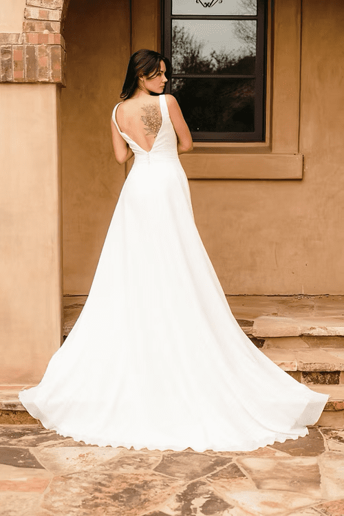1756319982668-0baeb4_ed1c81fa190b4f50a2e913fdb82e7045-mv2 Noemi, an ivory/ivory A-line wedding gown with a deep V-back, wide straps, and a soft, flowing chiffon chapel train. The bride, with dark hair and a floral tattoo visible on her upper back, stands with her back to the camera, looking over her left shoulder, on a stone patio against a warm-toned building.