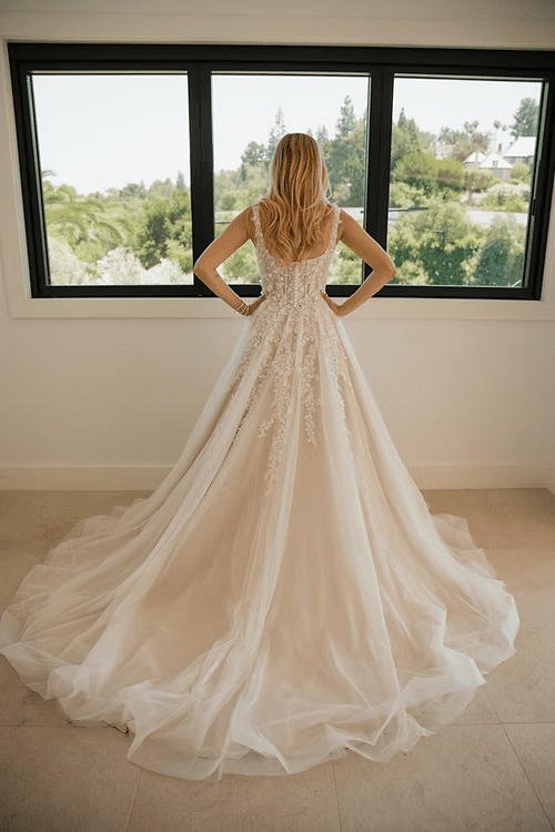 1756320379079-0baeb4_470aaba1399a465c81424f7147736a0f-mv2 Kinsley, an ivory over mocha ballgown bridal gown, is shown from behind on a blonde bride standing with her hands on her hips, gazing out a large window. The dress features a voluminous tulle skirt with a cathedral train, and a bodice and skirt adorned with intricate 3D embroidered lace with floral patterns and delicate embellishments. A scoop neckline is visible on the back. Outside the window, a sunny green landscape is visible.