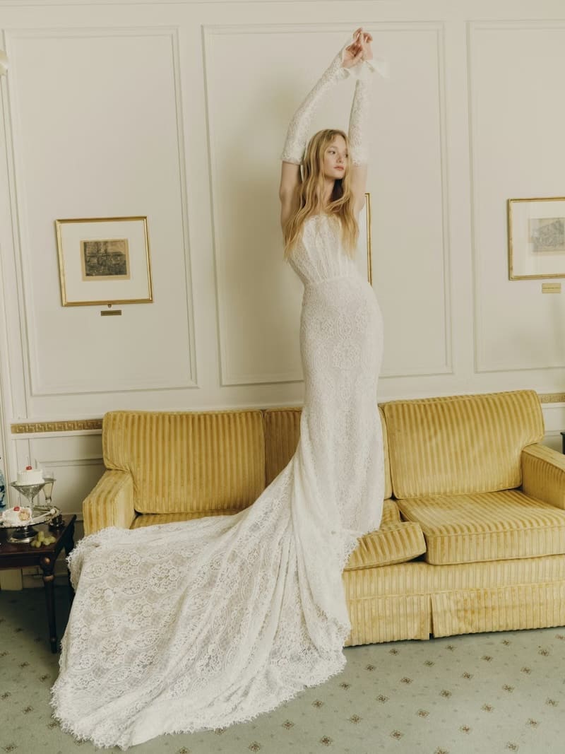 1765922143353-ss26-alyne-emme-3-front Emme, an ivory and nude strapless sheath wedding gown, made of delicate translucent lace with pleated tulle detailing at the neckline. The gown features a line of pearl buttons down the back and a sweeping cathedral train, completed with matching lace opera sleeves. A woman gracefully poses in the gown, standing on a yellow sofa in a grand room with white paneled walls and framed artwork, her arms reaching upwards.