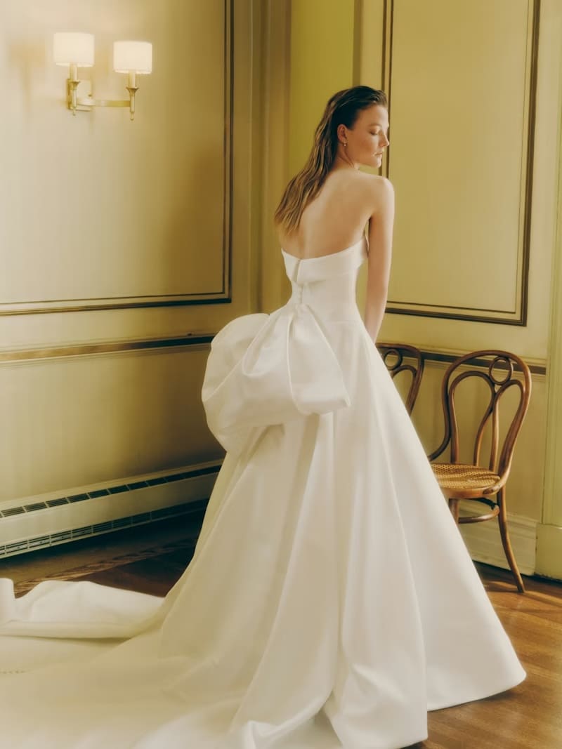 1765922544367-Z_fQB-vxEdbNO2qJ_RV_SS26_RIVINI_WELLS-1-BACK Wells, an ivory strapless Basque waist ballgown with a cuffed neckline and architectural bodice. Its voluminous skirt flows into a dramatic cathedral train, with a luxurious bow and button trail adorning the back. The gown is seen from behind on a model with wet-look hair, in an elegant, sunlit room.