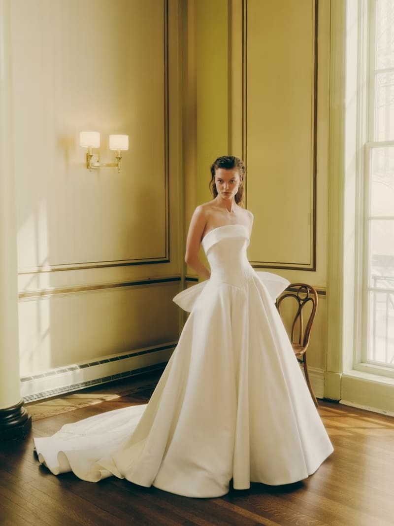 1765922544368-rivini-wells-ss26-2-front Wells, an ivory strapless satin basque waist ballgown featuring a refined cuffed neckline, an architectural bodice with precise seam detailing, a voluminous skirt, a luxurious bow at the back, and a dramatic cathedral train with a trail of buttons.