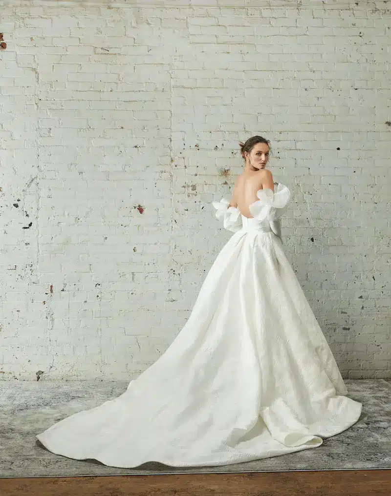FW22-LOOK-14-RIVINI-4707-XENIA-5642-v2-1 Xenia, an ivory strapless ballgown crafted from luxurious textured silk jacquard gazer, features a grand pleated skirt with a chapel-length train and an asymmetrically draped bodice. The gown is accessorized with whimsical detachable 3D petal sleeves. The model is captured from behind, looking over her shoulder, against a white brick wall.