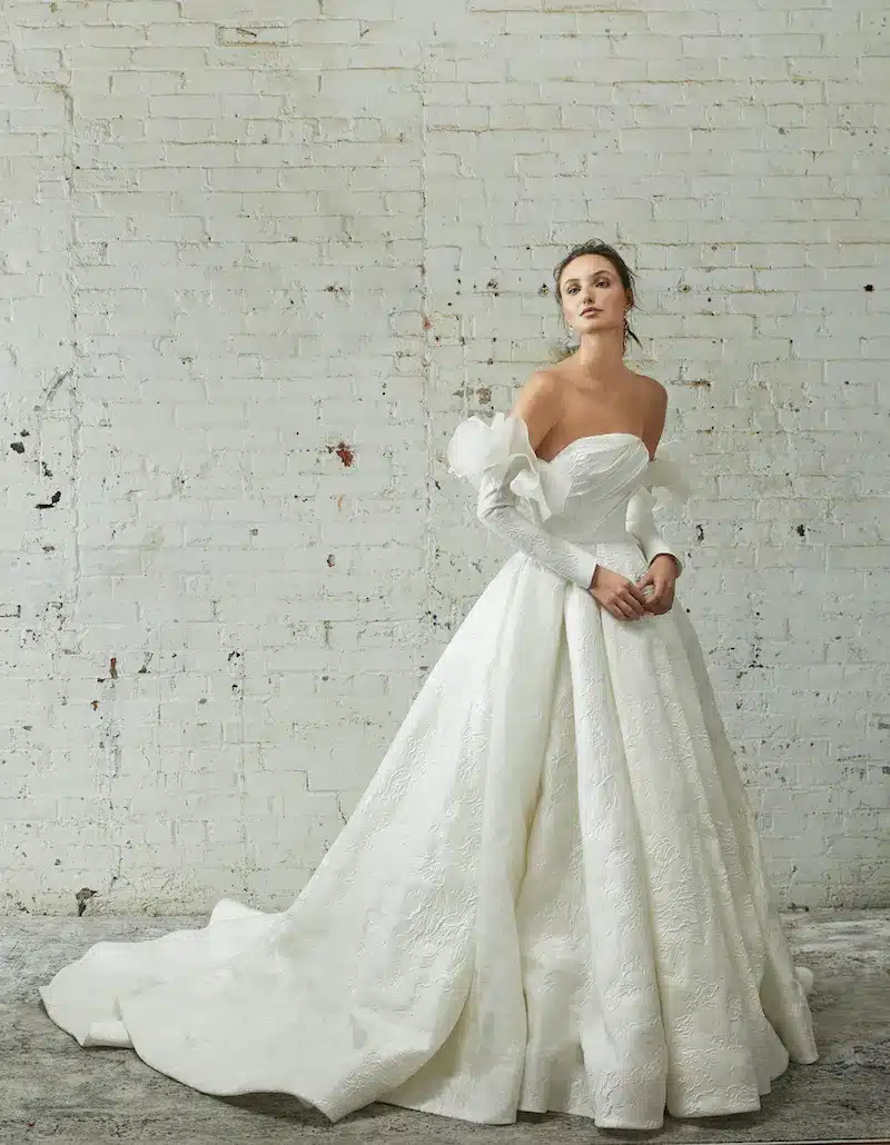 fw22-look-14-rivini-4707-xenia-5455-v3-3 Xenia, an ivory Rivini strapless ballgown, features a luxurious textured silk jacquard with a voluminous pleated skirt and an asymmetrically draped bodice. The gown is styled with detachable long 3D petal sleeves, and its chapel train trails elegantly on the floor.