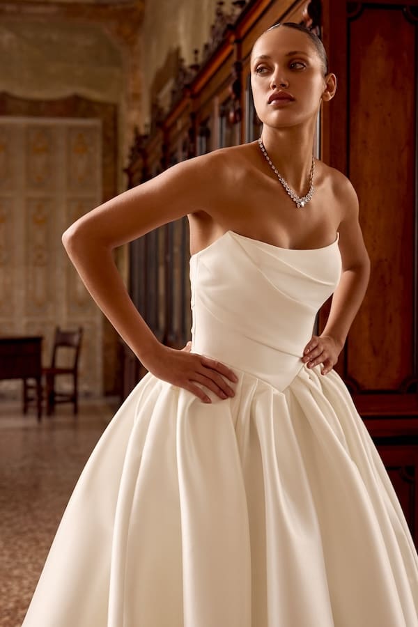 kelsey-fc-justin-alexander Kelsey, an ivory Mikado bridal ball gown by Justin Alexander, featuring a strapless scoop neckline, a delicately ruched bodice, a basque waistline leading to a voluminous skirt with knife pleating, hidden pockets, and a chapel-length train. The gown is worn by a model with sleek hair and a diamond necklace, posing with hands on her hips in an elegant, antique room.