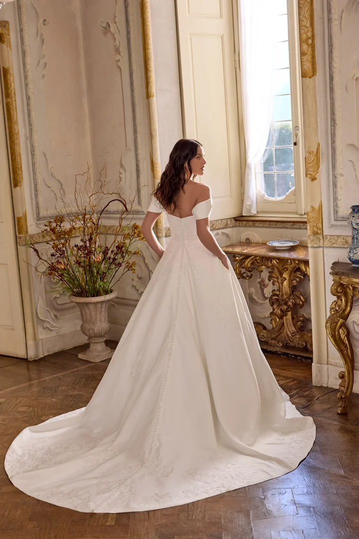 larissa-justin-alexander-3 Larissa, an ivory matte satin A-line wedding gown by Justin Alexander, shown from the back in an opulent room. The dress features a soft scoop neckline with detachable off-the-shoulder straps, a draped and ruched bodice, and tonal sequined lace appliqués cascading from the pleated bodice down the back. A row of buttons trails into the chapel-length train.