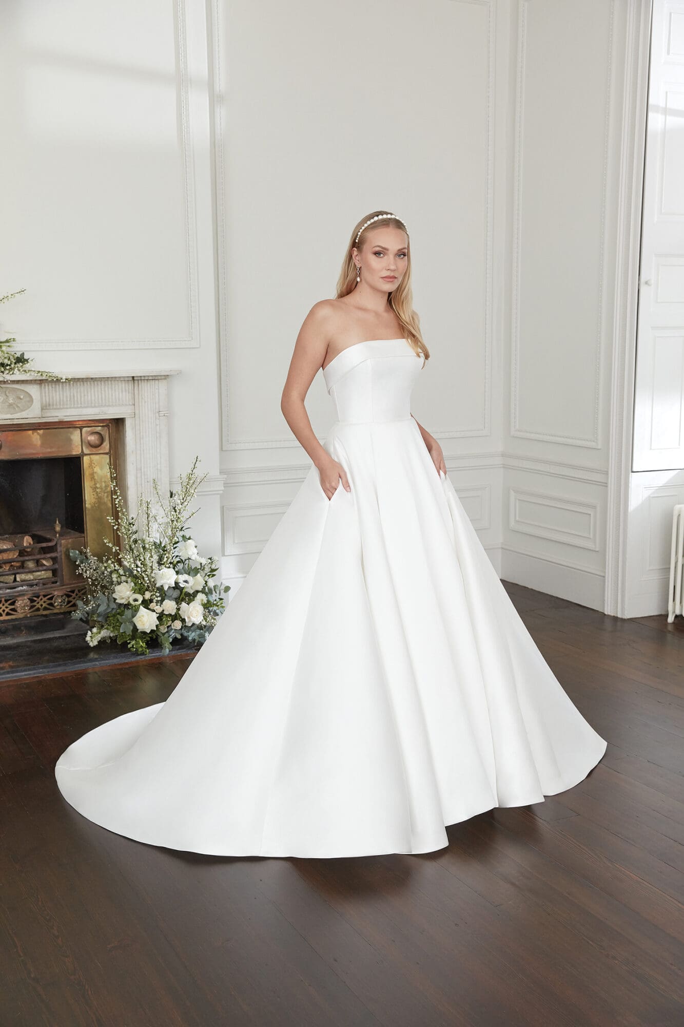 14-112397-1 A Sincerity by Justin Alexander strapless Mikado bridal ball gown with a chapel train. The elegant white dress features a structured bodice, a natural waistline, and a voluminous skirt with hidden pockets, as shown by the model's hands casually placed within them. The blonde model wears a pearl headband and drop earrings, standing in a classic room with a fireplace adorned with white flowers.