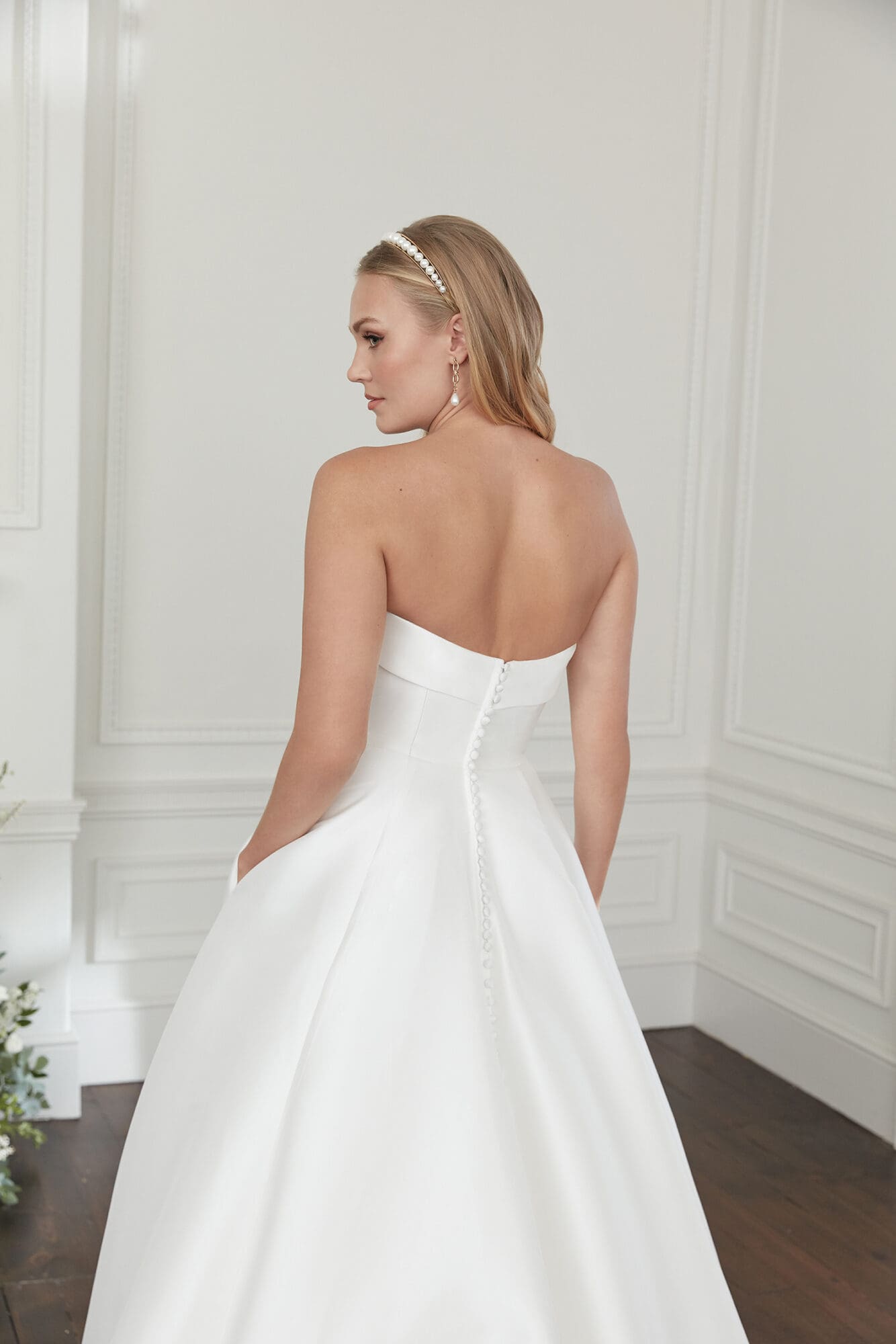 14_112397_2 A woman models an elegant white strapless Mikado ball gown by Sincerity by Justin Alexander, seen from the back as she looks over her left shoulder. The dress features a structured cuffed neckline, hidden pockets, and Mikado-covered buttons cascading down its chapel-length train. She accessorizes with a pearl headband and pearl drop earrings.