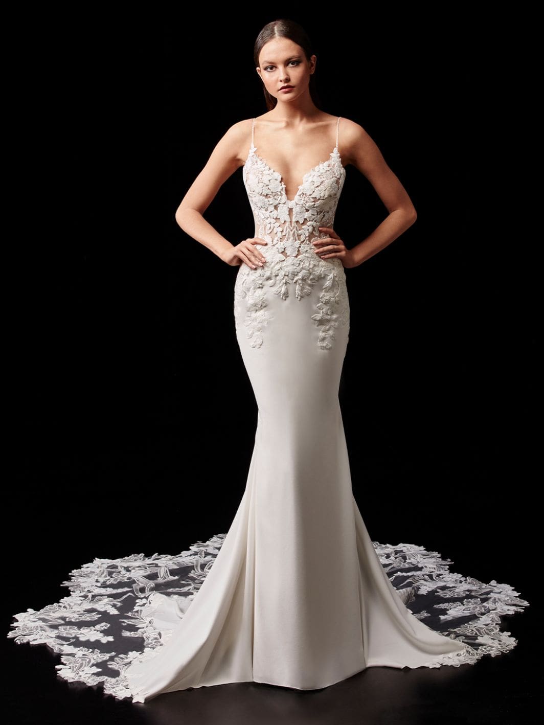 1598039410338-Enzoani_2021_Pro_Pearl_Fro_Web_1064_1418_80_int_s_c1 Pearl by Enzoani, an ivory and nude fit-and-flare bridal gown featuring a plunging illusion sweetheart neckline with delicate lace appliqués, spaghetti straps, and a low back. The elegant crepe skirt gracefully transitions into a dramatic cathedral-length train with an intricate removable lace overlay.