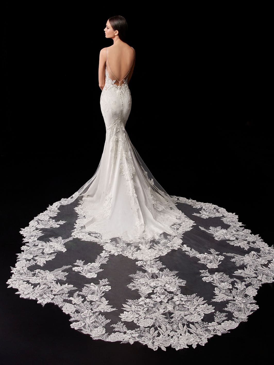 1598039547366-enzoani-2021-pro-pallas-bac-web-1064-1418-80-int-s-c1 Pallas, an Ivory/Nude fit-and-flare bridal gown with spaghetti straps, is shown from the back on a model looking over her left shoulder against a black background. The gown features a deep V-back with delicate beaded straps and a fitted bodice showcasing illusion and sheer elements with contemporary sequin floral embroidered lace. Made from luxurious stretch georgette, the dress hugs the figure before flaring into a dramatic cathedral-length train. The train is exquisitely detailed with intricate floral lace appliqué and embroidery on sheer tulle, spreading wide across the floor, creating a breathtaking and elegant silhouette.