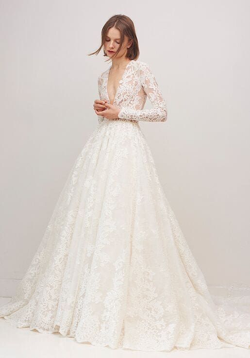 1603133708318-5ce0f326-7bd1-438c-a779-d8331ba266d3-rs-515-h-2 Biltmore, an ivory Rivini bridal ball gown featuring a deep V-neckline, long lace sleeves, and an intricate lace overlay throughout its voluminous skirt and cathedral train, modeled by a woman with short brown hair.