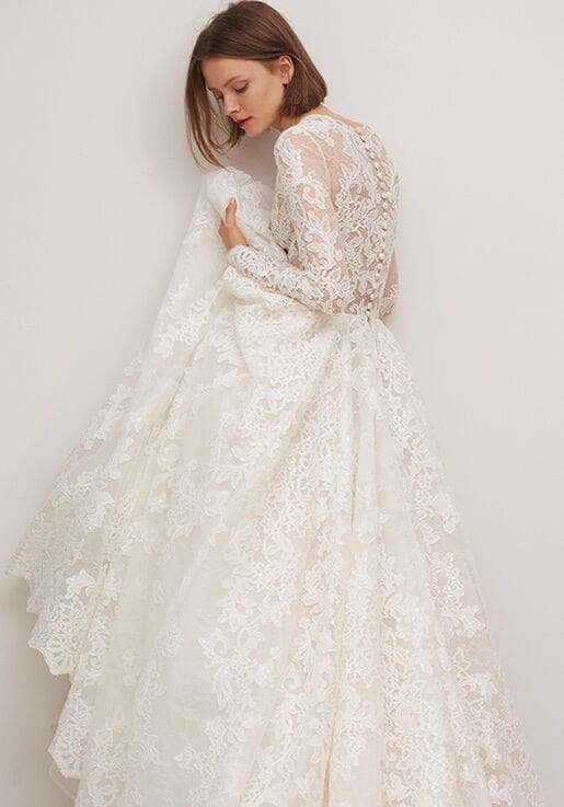 1603133708319-c10798a4-8dee-4b91-b338-a50ae6217af4 Biltmore, an ivory lace long-sleeved bridal ballgown by Rivini, featuring a deep V-neck and delicate buttons cascading down the back to a sweeping cathedral train. The model is shown from a side-back angle, looking down, holding up the voluminous skirt to highlight the intricate lace details.