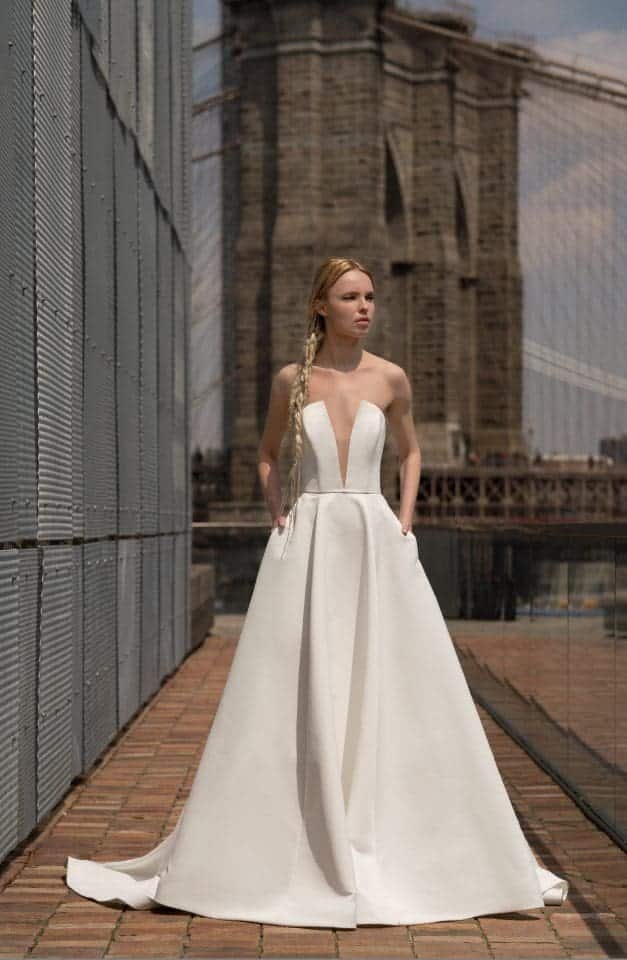Blanchett, an ivory Alyne bridal gown with a strapless plunge V-neckline, an A-line satin skirt, and pockets, worn by a blonde model with a long braid. She stands on a brick path alongside a metal wall, with a historic stone bridge in the sunny background.